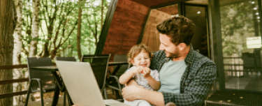 A father holding his young while sitting outdoors in front of his computer. 