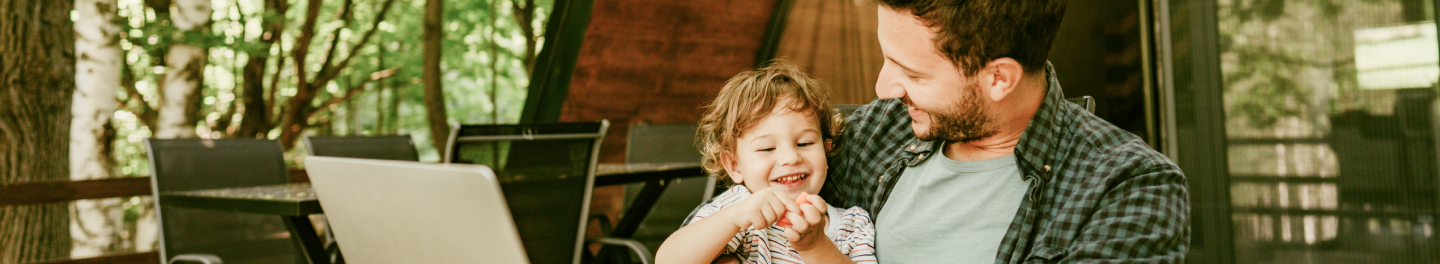 A father holding his young while sitting outdoors in front of his computer. 